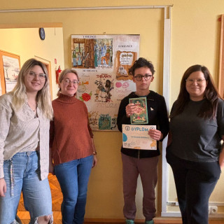 4 osoby stoją na tle plakatu prac nagrodzonych w konkursie