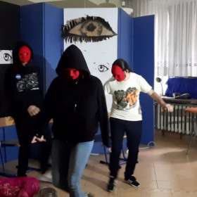 Dziewczyna leży na podłodze, wokół niej krążą osoby w maskach