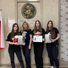 Trzy uczennice wraz z nauczycielem przed tablicą Józefa Piłsudskiego