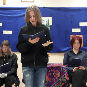 Dwie osoby stoją i dwie osoby siedzą, jedna osoba czyta wiersz.