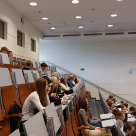 Uczniowie siedzący w auli słuchają referatu.
