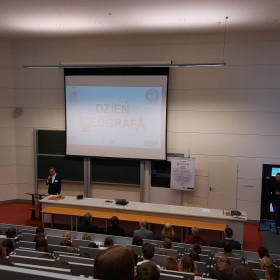 Pracownik naukowy instytutu prezentuje program obchodów przemawiając na auli.