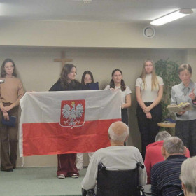 Na scenie dziewczyna trzyma flagę, nauczyciel recytuje wiersz.
