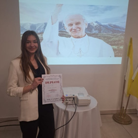 Oliwia z dyplomem w ręce na tle obrazu Jana Pawła II.