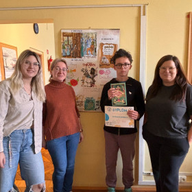 4 osoby stoją na tle plakatu prac nagrodzonych w konkursie