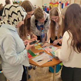 Grupa uczniów pochyla się nad okrągłym stolikiem, na którym leżą paski kolorowego papieru. Uczniowie rozwiązują rebus. Wolontariuszka z Azerbejdżanu kontroluje na telefonie czas pracy uczniów.