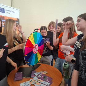 Grupa uczniów stoi przy stanowisku z kolorowym kołem do gry. Obracają kołem, losując zadanie.